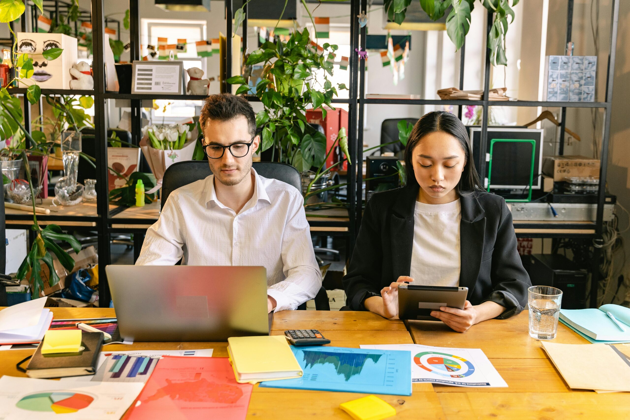 Two professionals working with laptops and tablets in a modern coworking space surrounded by plants.